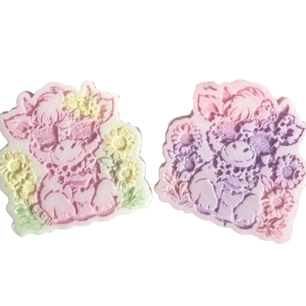 Two pastel-colored decorative highland cow shaped items with floral accents, shown on a plain white background. wax melt