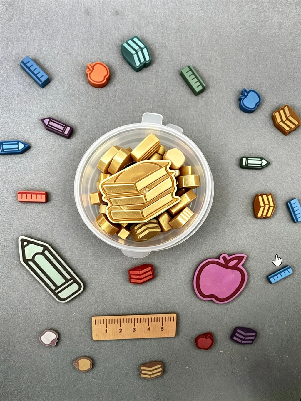 A clear plastic container filled with small gold-colored school-themed wax melts, surrounded by scattered matching charms in blue, green, pink, red, and beige on a gray background.