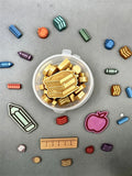 A clear plastic container filled with small gold-colored school-themed wax melts, surrounded by scattered matching charms in blue, green, pink, red, and beige on a gray background.
