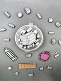 A collection of silver metal school-themed scoopies wax melts, including books, rulers, pencils, and apples, arranged around a clear plastic storage container on a gray background.