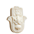 An off-white Hamsa hand soap bar featuring embossed details of a lotus flower, an eye of Providence, and intricate scrollwork, shown on a plain white background. CANDLE