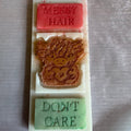 A rectangular white mold or tray holding three carved swax snap bar: a coral pink bar reading 'MESSY HAIR,' a translucent amber-brown bar with an embossed monkey face, and a pale green bar reading 'DON'T CARE,' all placed on a gray textured fabric background.