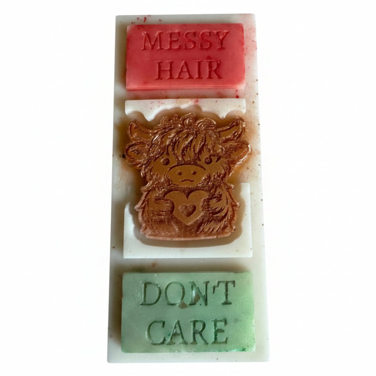 A rectangular white wax bar with three stacked colored sections: a pink top section embossed with 'MESSY HAIR', a brown cow-face center section, and a light green bottom section embossed with 'DON'T CARE'.