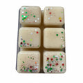 Six square cream-colored resin cubes with embedded red, green, silver, and white glitter, arranged in a clear plastic tray on a gray textured surface.