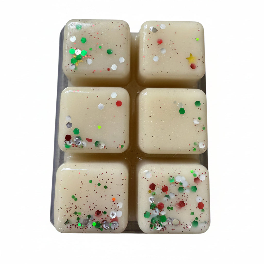 Six square cream-colored resin cubes with embedded red, green, silver, and white glitter, arranged in a clear plastic tray on a gray textured surface.