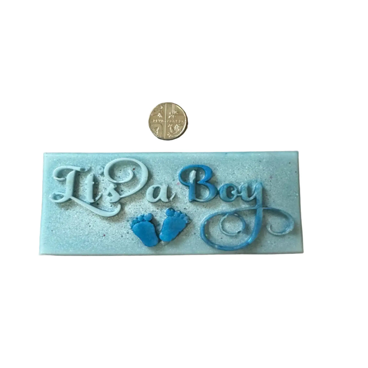 A rectangular light blue decorative plaque with raised script reading 'It's a Boy,' two small blue baby footprints, and a gold coin placed above it for scale, on a plain white background. ax melt