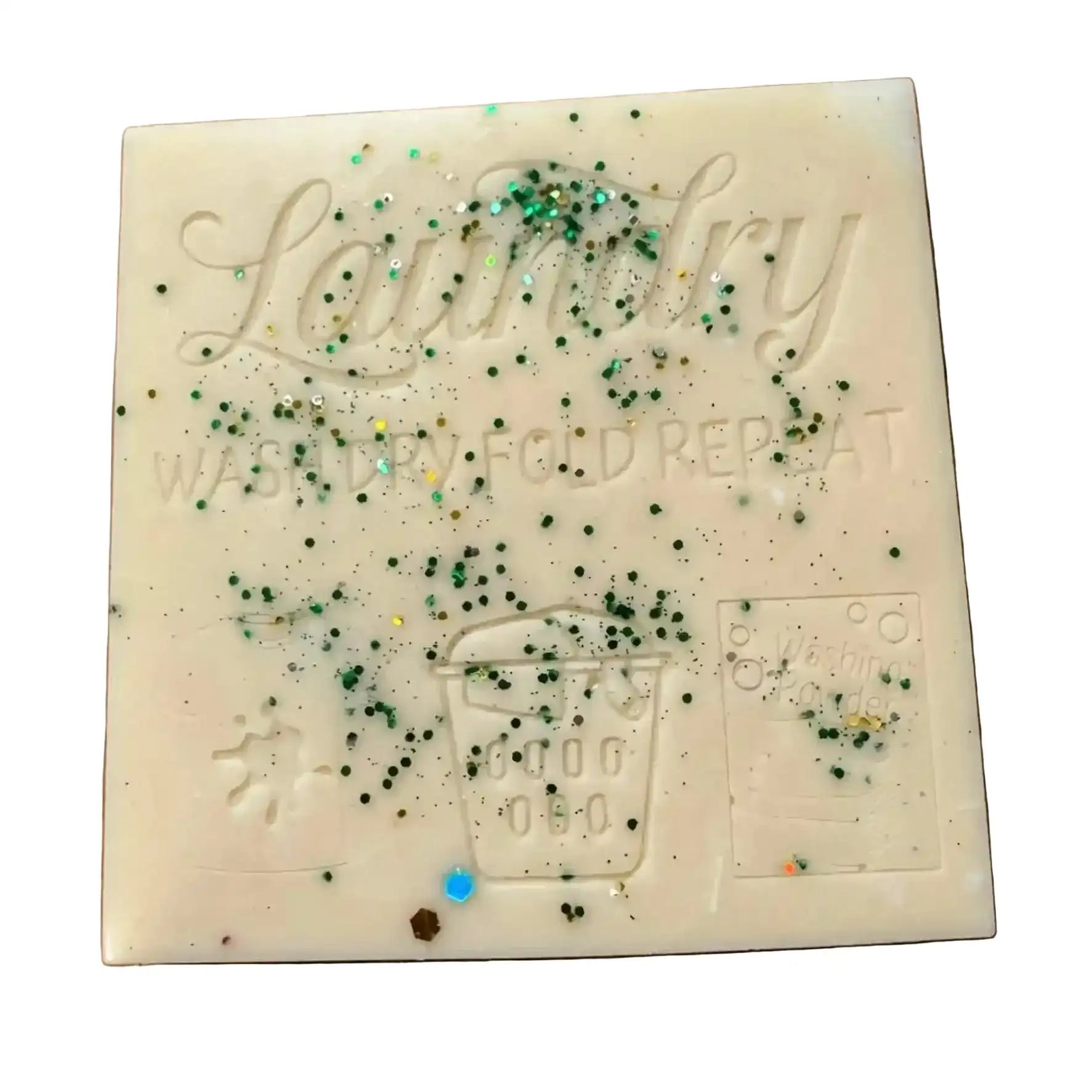 A square cream-colored soap bar embossed with 'Laundry' and 'WASH DRY FOLD REPEAT,' decorated with scattered green and blue glitter, on a plain white background. smash plate wax melt