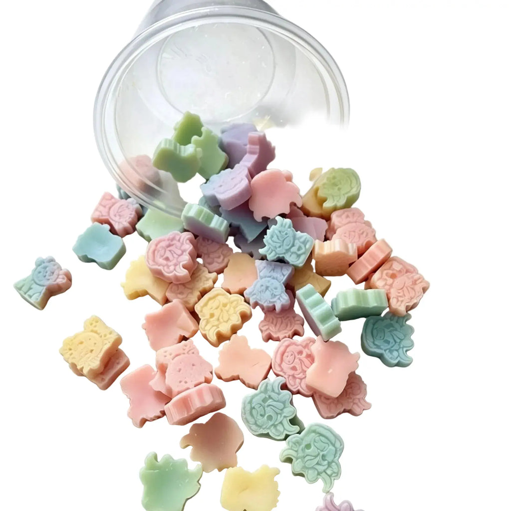 A clear plastic cup tipped over on a white background, spilling a mix of small pastel animal-shaped candies or soap pieces in pink, green, yellow, blue, and purple. WAX MELT SCOOPIES