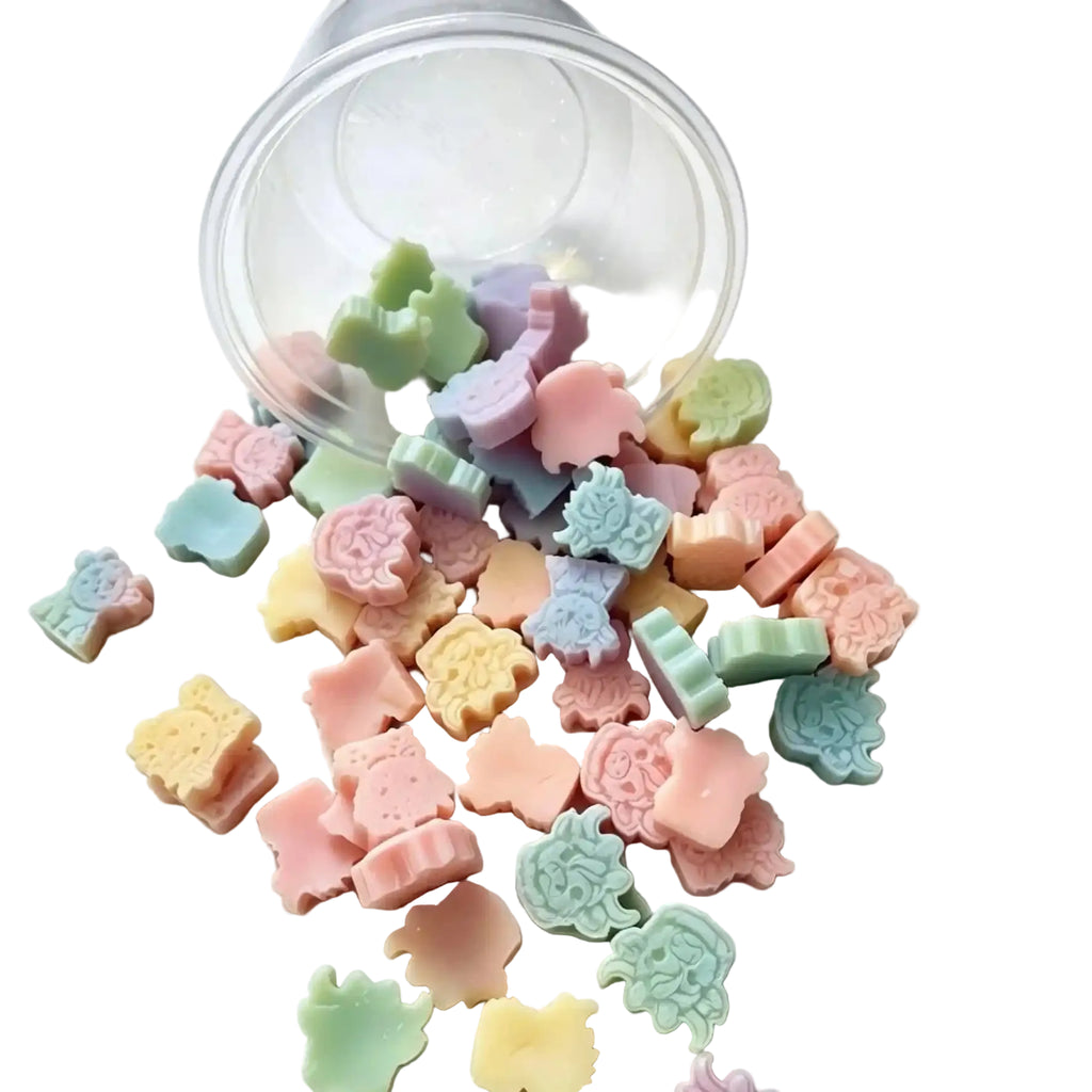 A clear plastic cup tipped over on a white surface, with many small pastel-colored, animal-shaped candy-like pieces spilled out. The pieces are in soft pink, peach, yellow, mint green, and light blue tones. WAX MELT