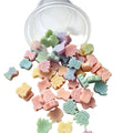 A clear plastic cup tipped over on a white surface, with many small pastel-colored, animal-shaped candy-like pieces spilled out. The pieces are in soft pink, peach, yellow, mint green, and light blue tones. WAX MELT