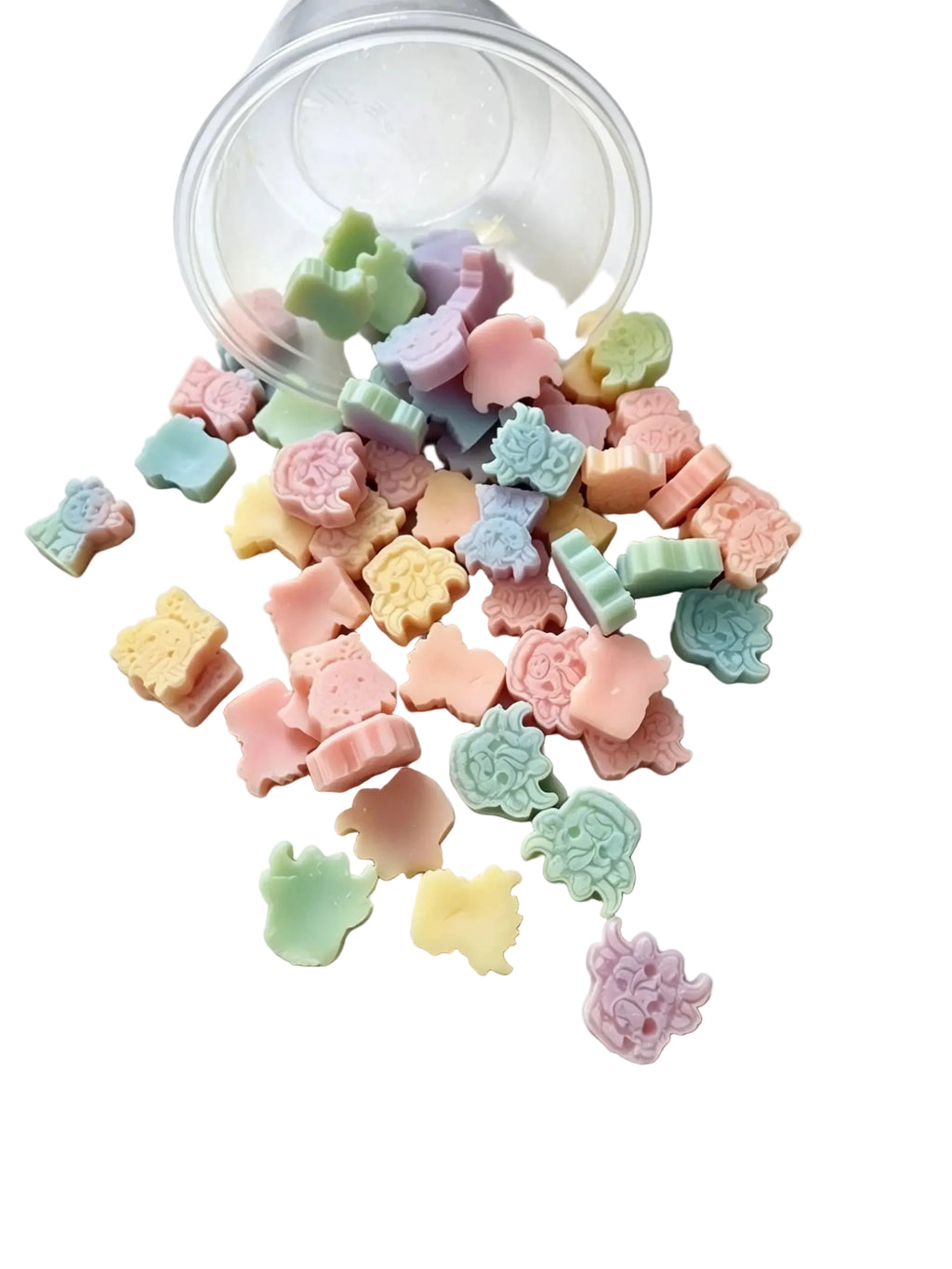 A clear plastic cup tipped over on a white surface, with many small pastel candy-like pieces spilled out in front. The pieces are shaped like animals and flowers in soft pastel colors including pink, blue, green, yellow, and lavender. scoopies wax melts 