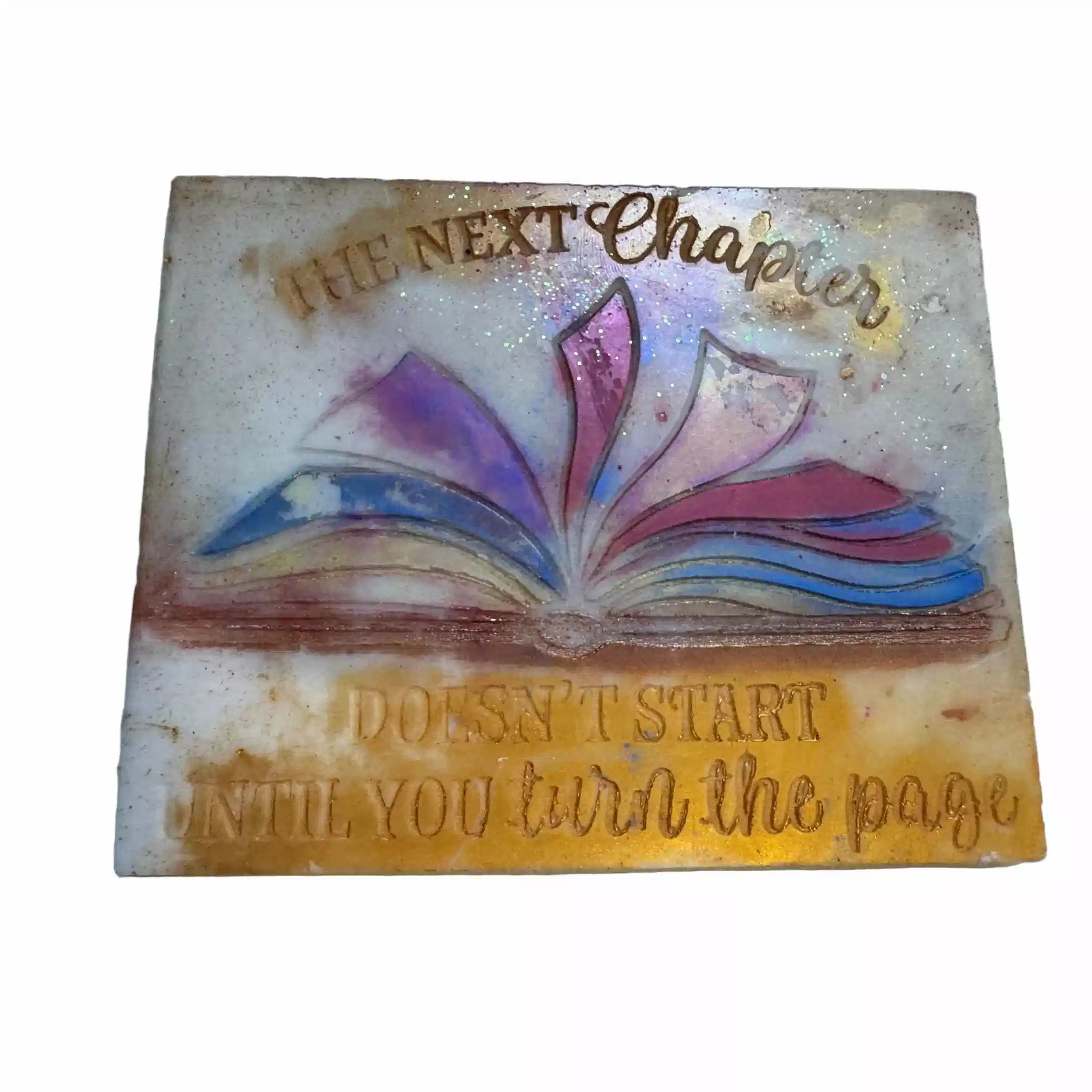 A square decorative book-themed tile or plaque with embossed text reading 'THE NEXT Chapter' and 'DOESN'T START UNTIL YOU turn the page,' surrounding an open book illustration with purple, pink, blue, and yellow tones on a light background. wax melt smash plate