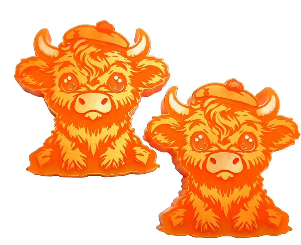 Two bright orange cartoon Highland cow wax melts with small orange hats, shown on a plain white background. One cow is facing forward and the other is turned to the side, both with large expressive eyes and textured fur details.