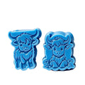Two blue cow-shaped wax melts wax melts on a white background, with black text above stating dimensions and wax weight. The left item is a highland cow facing left, and the right item is a smiling highland cow facing forward with small flowers at the base.