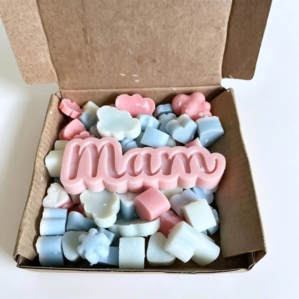 A cardboard gift box filled with pastel wax melts or soap pieces, including small cloud, flower, and star shapes in pink, light blue, and white, with a large pink 'Mam' word piece on top. scoopies wax melts
