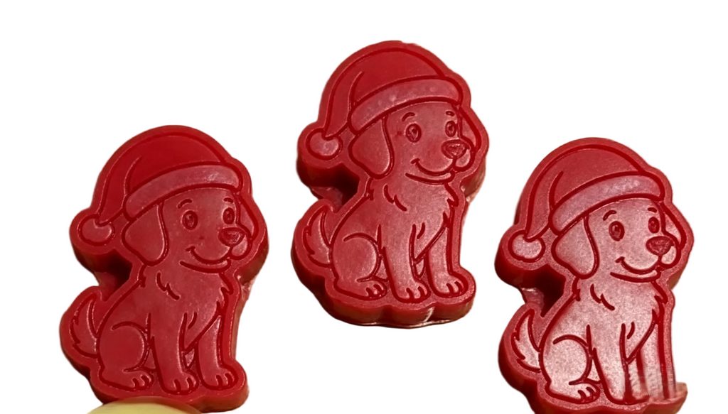 Three red dog-shaped wax melts wearing Santa hats arranged on a light wood grain surface.